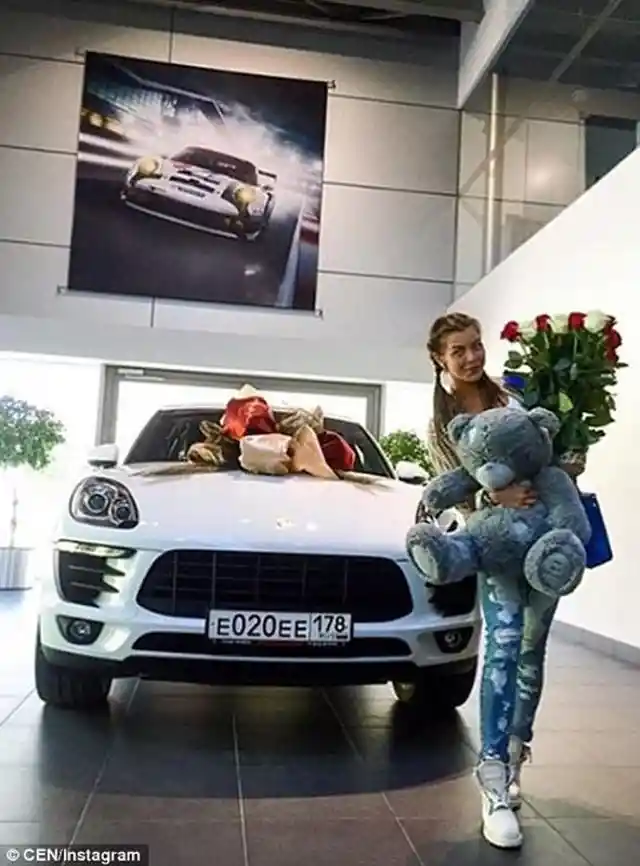 A young girl stands next to a brand new Porsche with flowers and a teddybear